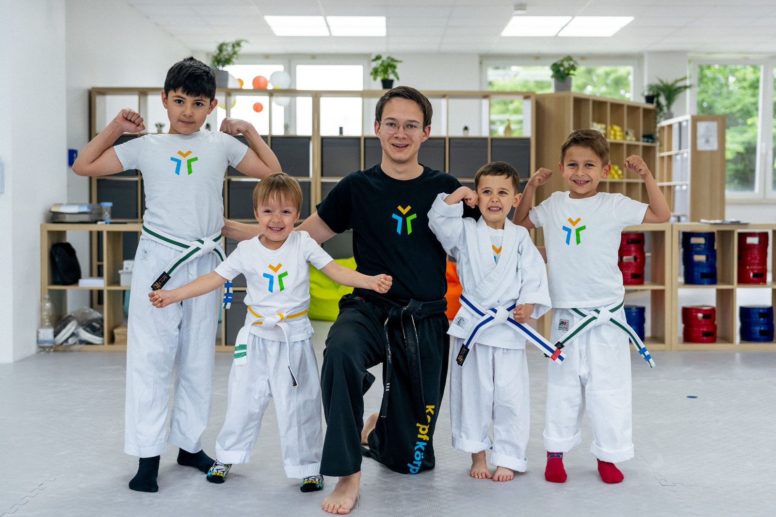 Trainer mit vier Kindern vor der Regalwand, alle zeigen ihre Muskeln